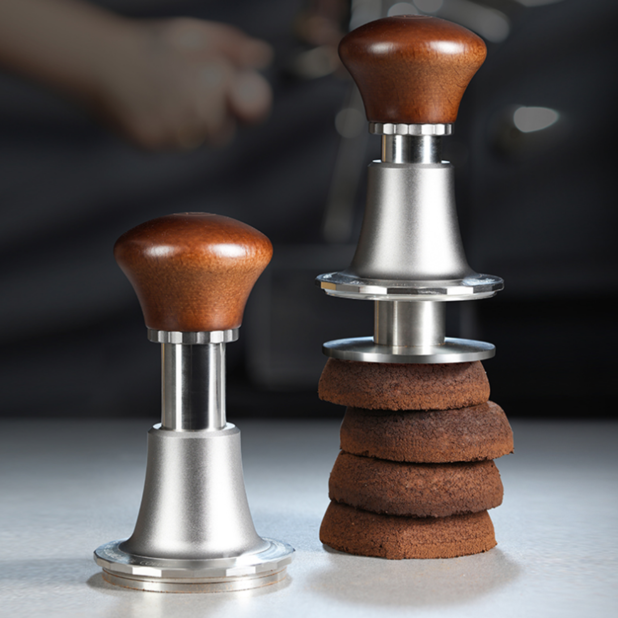 Adjustable espresso tamper with ergonomic wooden handle and stainless steel base, demonstrating even coffee puck compression with stacked used coffee pucks.