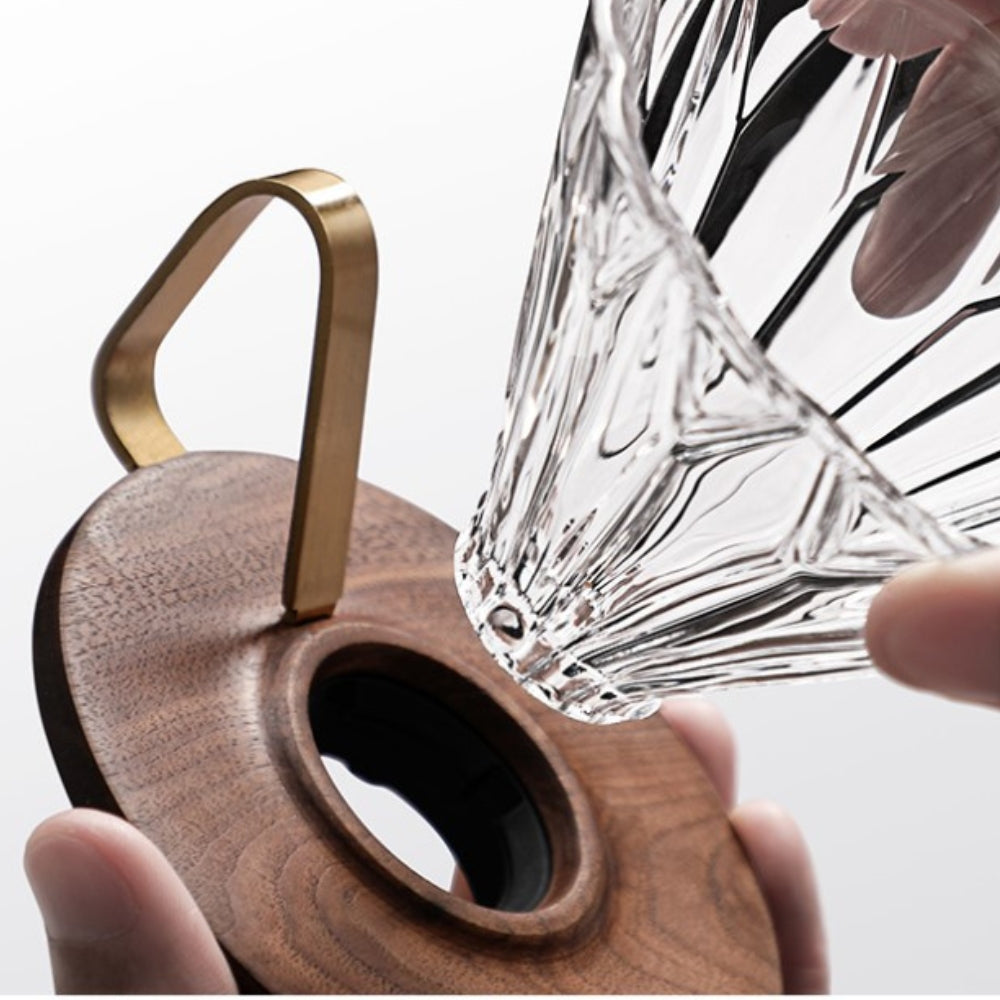 LAMOSE zeroHero Walnut Prisma Dripper. Close-up of a hand placing a clear textured glass dripper onto a dark walnut wood base with a brass handle.